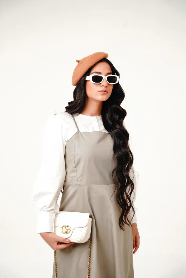 A woman wearing a beige pinafore dress with a white top, featuring a vintage-inspired neckline and wide straps.